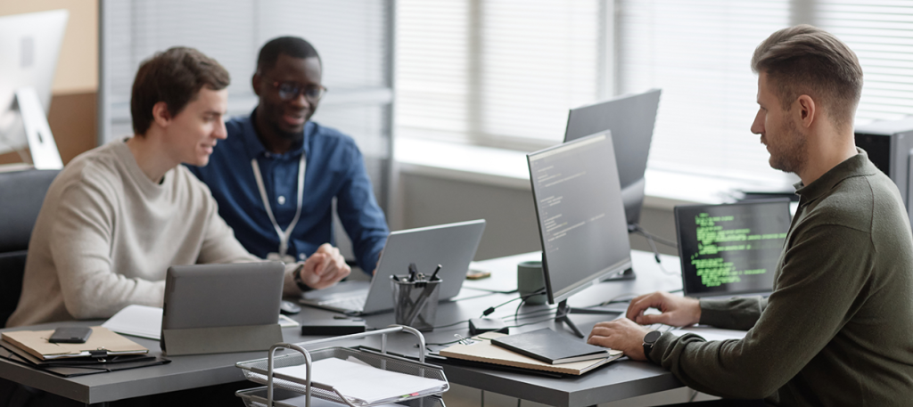 Three developers collaborate at a desk with laptops and code visible on monitors in a bright office.