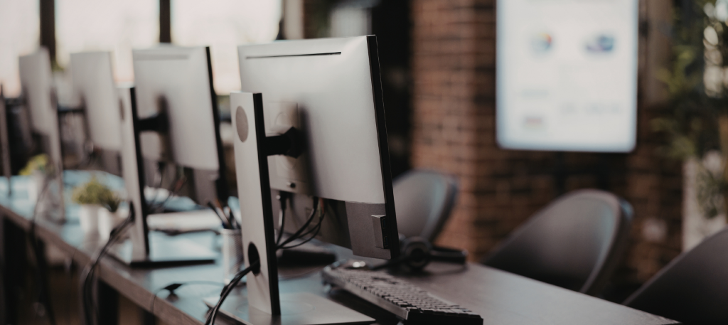 Row of computer monitors on a long desk in a modern office, with keyboards and cables visible.