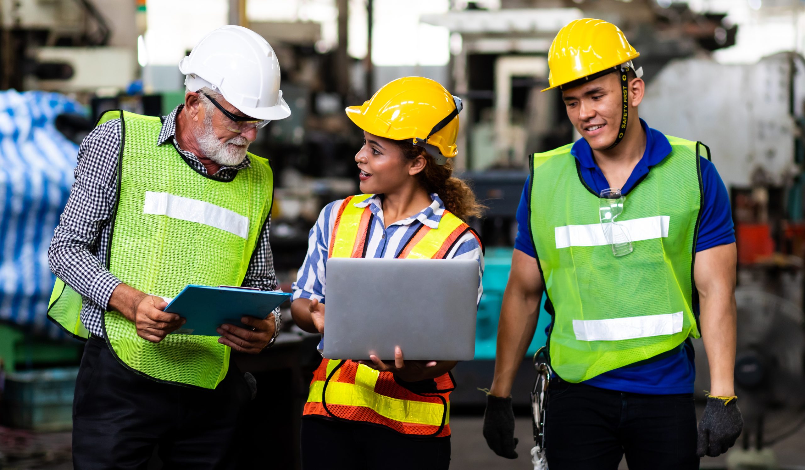 Three engineers in hard hats and safety vests reviewing a laptop and tablet on a factory floor